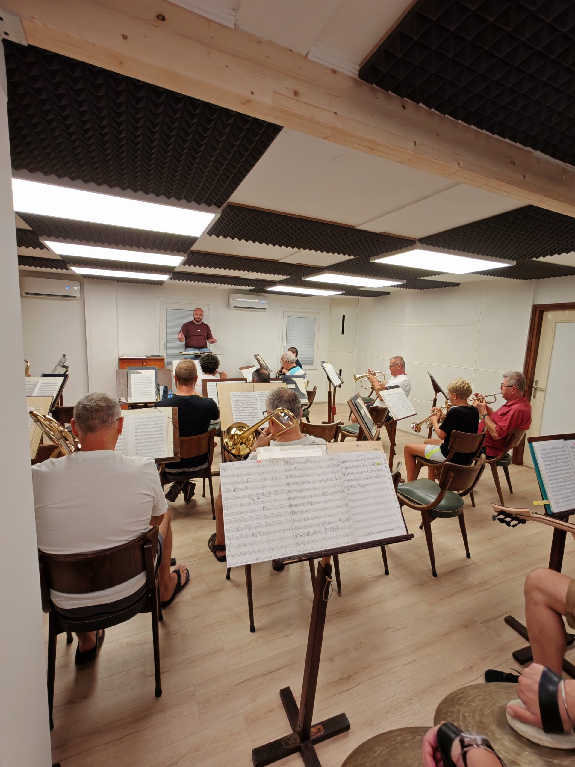 Puhački orkestar na probi u novoj dvorani, dirigent ispred orkestra, drvene grede i akustične ploče na stropu